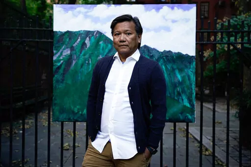 Bobby Calvan poses in front of a painting of the view from his childhood home in Waimanalo, Hawaii, Sunday, Oct. 9, 2022, in New York. His journey out of the closet has taken decades. Now 57, he is still sharing his truth about his sexuality. The journey hasn’t ended; indeed, it’s just beginning as he now lives his life as the man he believes he was meant to be. (AP Photo/Julia Nikhinson)