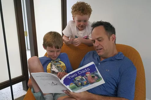 Wes Brown, right, reads to children Shawn Larimer-Brown, 7, left, and Charlie Larimer-Brown, 5, center, at their home Friday, Aug. 18, 2023, in Winter Park, Fla. Across the country, books and lessons that represent different families and identities to the youngest of learners are increasingly the target of the conservative pushback to efforts to promote diversity and inclusion. (AP Photo/John Raoux)