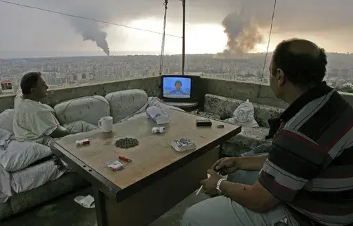 Lebanese citizens watch Hezbollah leader Sheik Hassan Nasrallah speak on television as black smoke rises following Israeli attacks on a Hezbollah stronghold in the southern suburbs of Beirut, Lebanon, on July 16, 2006. (AP Photo/Kevork Djansezian, File)