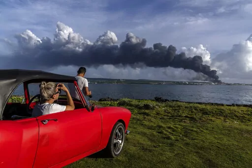 People watch a huge plume of smoke rise from the Matanzas supertanker base, as firefighters work to douse a fire that started during a thunderstorm the night before, in Matanzas, Cuba, Sunday, Aug. 7, 2022. Cuban authorities say lightning struck a crude oil storage tank at the base, sparking a fire that sparked four explosions that injured more than 121 people, one person dead and 17 missing. (AP Photo/Ramon Espinosa)