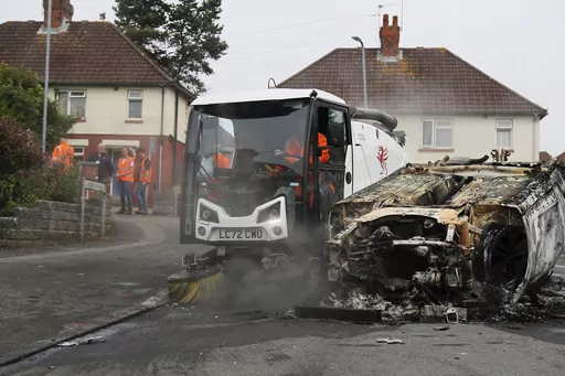Council workers clear debris from the area immediately around a car that was set alight in Ely, Cardiff, Tuesday, May 23, 2023. Several dozen youths pelted police with objects and set cars ablaze on Monday evening in Cardiff in local unrest that erupted after two teenagers died in a road accident. (PA via AP)