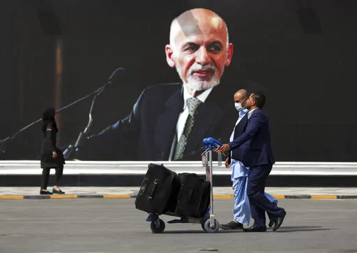 Passengers walk to the departures terminal of Hamid Karzai International Airport in Kabul, Afghanistan, on Saturday, Aug. 14, 2021, past a mural of President Ashraf Ghani, as the Taliban offensive encircled the capital. On the eve of the anniversary of the Taliban takeover of Kabul, Afghanistan's former president on Sunday, Aug. 14, 2022, defended what he said was a split-second decision to flee, saying he wanted to avoid the humiliation of surrender to the insurgents. (AP Photo/Rahmat Gul, File