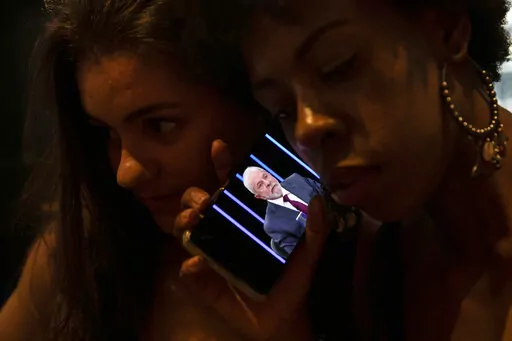 Women listen on a mobile phone to an interview of former president Luiz Inacio Lula da Silva, who is running again for president, with Jornal Nacional on TV Globo, outside a bar in Rio de Janeiro, Brazil, Thursday, August 25, 2022. The Superior Electoral Court, the country's top electoral authority, announced Thursday, Oct, 20, that it would be banning "false or seriously decontextualized" content that “affects the integrity of the electoral process.” No request from a prosecutor or complain