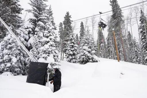 Ian Sidwell adjusts a machine used to make snow at Vail Mountain Resort as snowboarders ride a lift, Wednesday, Dec. 29, 2021, in Vail, Colo. Newer snowmaking technology is allowing ski areas to be more efficient with energy and water usage as climate change continues to threaten snowpack levels. (AP Photo/Brittany Peterson)