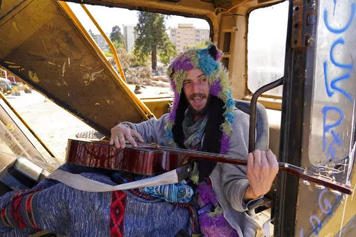 At Atmaa Das, 28, who is homeless from Alabama, strums his guitar while singing parts of Woody Guthrie's anthem, "This Land Is Your Land" while sitting in a piece of construction equipment left behind at People's Park in Berkeley, Calif., on Aug. 16, 2022. The three-acre site's colorful history, forged from University of California, Berkeley's seizure of the land in 1968, has been thrust back into the spotlight by the school's renewed effort to pave over People's Park as part of a $312 million p