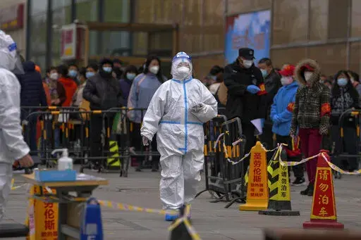 A health worker wearing a protective suit walks by masked residents who wait in line to get their throat swab at a coronavirus testing site following a COVID-19 case was detected in a residential buildings, Wednesday, April 6, 2022, in Beijing. (AP Photo/Andy Wong)