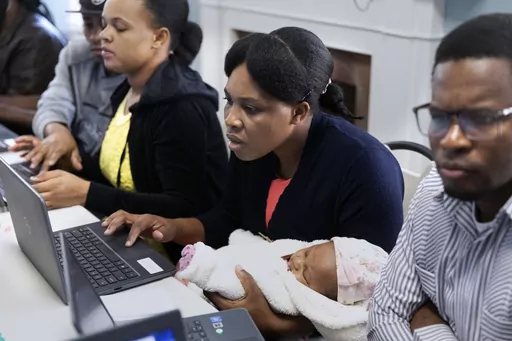 Jimene Admettre, center, sits next to her husband Ernseau, right, and holds her daughter Gabyana while learning computer skills, Friday, Dec. 22, 2023, in a rectory building where they are staying at the Bethel AME Church in the Jamaica Plain neighborhood of Boston. Demand for shelter has increased as Massachusetts struggles to find newly arriving migrants places to stay after hitting a state-imposed limit of 7,500 families in its emergency homeless shelter system last month. (AP Photo/Michael D