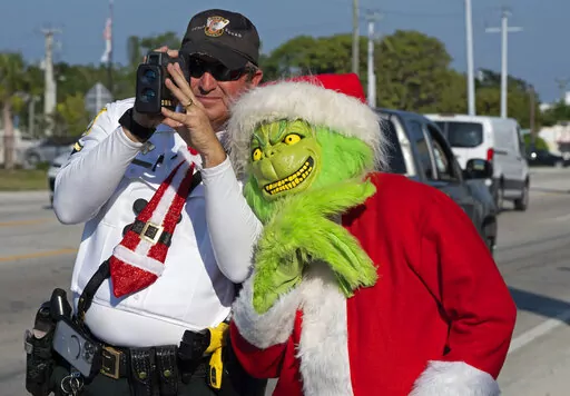 In this photo provided by the Florida Keys News Bureau, Monroe County Sheriff's Office Colonel Lou Caputo, right, costumed as the Grinch, leans on the shoulder of Deputy Andrew Leird, left, as he uses a laser speed detector to check speeds of motorists traveling through a school zone on the Florida Keys Overseas Highway Tuesday, Dec. 13, 2022, in Marathon, Fla. For drivers slightly speeding through the area, Caputo offers them the choice between an onion or a traffic citation. (Andy Newman/Flori
